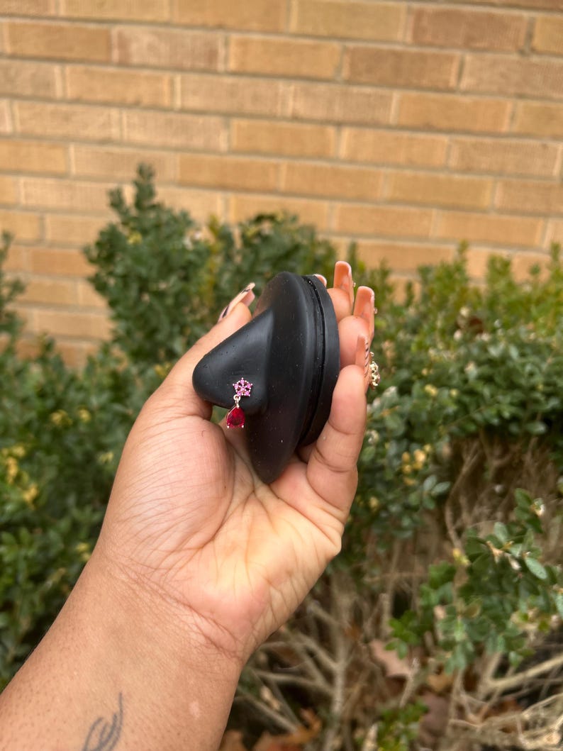 May include: A black nose-shaped display stand holding a small, dangling earring. The earring has a pink floral design and a teardrop-shaped red gem. The background features a brick wall and green foliage.