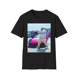 Może przedstawiać: Czarny t-shirt z nadrukiem przedstawiającym mężczyznę pozującego przed różowym samochodem sportowym. W samochodzie są jeszcze dwóch mężczyzn. Mężczyzna z przodu ma na sobie błękitną koszulę i beżowe spodnie.