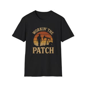 Op de afbeelding: Zwart t-shirt met een grafisch ontwerp met een silhouet van een persoon die naar een oliepomp loopt, tegen een zonsondergang. De tekst "Workin' the Patch" is boven en onder de afbeelding gebogen.
