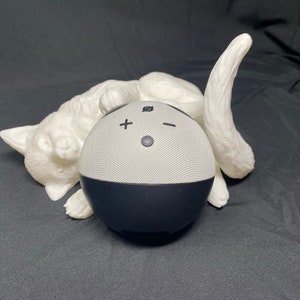 May include: A white ceramic sculpture of a cat with a black and white smart speaker resting on its back.