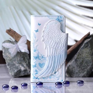May include: White wallet with a blue angel wing design. The wallet features a heart and butterfly motif, with blue accents. The design is embossed on the front of the wallet. The wallet is rectangular and has a snap closure.