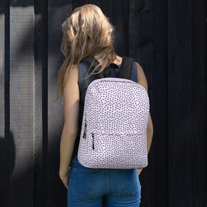 May include: A white backpack patterned with small, dark purple shapes. The backpack features black straps and a black zip. The person in the image is wearing blue jeans and a grey top.