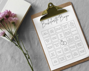 May include: A clipboard with a printed "Bachelorette Bingo" game sheet. The sheet has various squares with activities. A sprig of pink flowers and a closed book are on the left.