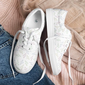 May include: White canvas sneakers with a repeating pattern of colorful lightning bolts. The shoes have white laces and soles. The sneakers are displayed on a pink and tan fabric background.