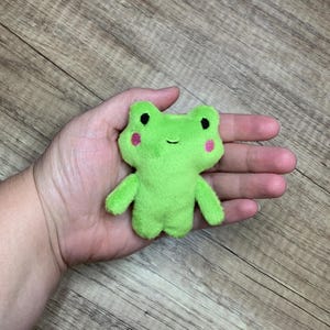 May include: A small, bright green plush frog toy with a soft, fuzzy texture. The frog features two black embroidered eyes, a simple stitched mouth, and two small pink embroidered circles on its cheeks, giving it a friendly and simple appearance.