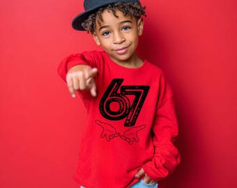 May include: A red long-sleeve shirt with the number "67" in black, along with two hand gestures. The shirt is worn by a person wearing a black baseball cap. The background is a solid red.
