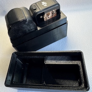 Puede incluir: Accesorios de cámara negros impresos en 3D, que incluyen una cámara, un micrófono y una funda protectora. La cámara está montada sobre una base negra. El micrófono tiene forma redondeada negra. La funda está abierta y vacía.