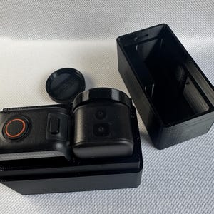 Puede incluir: Cámara de acción negra con un botón rojo y una tapa de lente, sobre una base negra. Una funda negra separada está cerca. La cámara está diseñada para capturar videos y fotos.