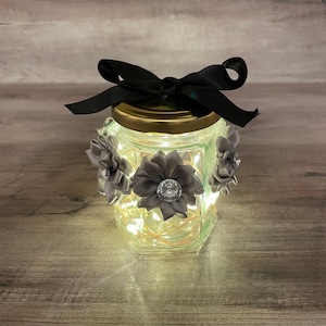 Pode incluir: Um frasco de vidro com tampa dourada e um laço de fita preto. O frasco é decorado com flores de tecido cinza e um pequeno strass. O frasco é iluminado por dentro com luzes brancas de fadas.