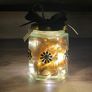 May include: A glass jar with a black ribbon and gold accents. The jar is filled with gold-colored candy and has a string of lights inside. The jar is lit up and the lights are visible through the glass.