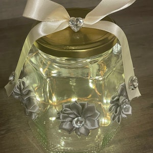 May include: A glass jar with a gold lid and a white satin ribbon tied around the top. The jar is decorated with gray satin bows and rhinestones. The jar is lit from within with white fairy lights.