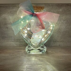 May include: A heart-shaped glass jar with a cork lid and a string of white lights inside. The jar is decorated with a pink and teal ribbon.