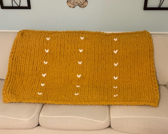 May include: A mustard yellow knitted throw blanket with white heart accents. The blanket is laying on a white couch.