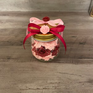 May include: A glass jar filled with pink and red fabric roses, tied with a pink and red ribbon, and topped with a gold lid.