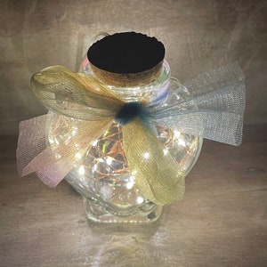May include: A heart-shaped glass jar with a cork lid and a blue and yellow ribbon. The jar is filled with white fairy lights.