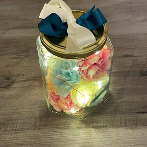 May include: A glass jar with a gold lid and a white and navy blue bow. The jar is filled with colorful artificial flowers and white fairy lights.