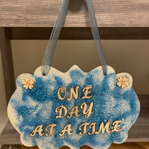 May include: A blue and white glitter sign with the words "ONE DAY AT A TIME" in gold letters. The sign is hanging from a gray ribbon.