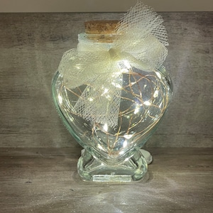 Peut inclure: Un bocal en verre en forme de cœur avec un couvercle en liège et un nœud en tulle blanc. Le bocal est rempli de lumières féériques blanches.