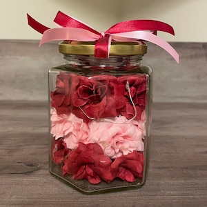 May include: A glass jar with a gold lid and a pink and red ribbon tied around the top. The jar is filled with three layers of fabric flowers, red, pink, and red.