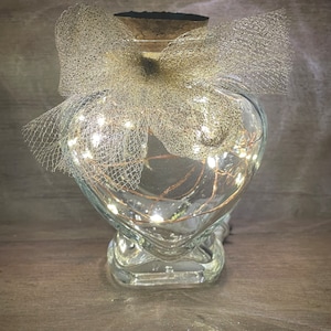 May include: A clear glass heart-shaped jar with a cork lid and a silver tulle bow. The jar contains a string of white fairy lights.