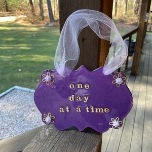 May include: Purple wooden sign with the text "one day at a time" in gold lettering. The sign is decorated with white flowers and has a white tulle bow.