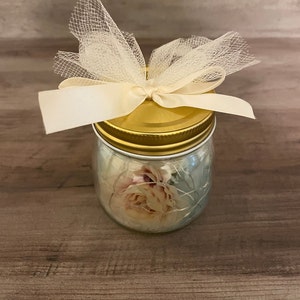 Op de afbeelding: Een glazen pot met een gouden deksel en een witte tule strik. De pot heeft een bloemmotief met een roze roos.