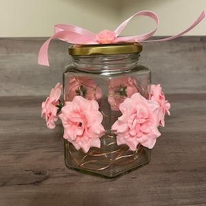May include: A clear glass jar with a gold lid and a pink ribbon tied around the top. The jar is decorated with pink flowers and a string of lights.