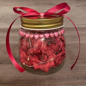 May include: A glass jar with a gold lid and a red ribbon tied around the top. The jar is filled with red rose petals and pink heart-shaped beads.