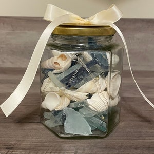 May include: A glass jar with a gold lid and a white ribbon tied around the top. The jar is filled with blue and white sea glass and seashells.