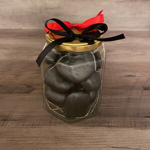 May include: A glass jar with a gold lid and a black ribbon tied around the top. The jar is filled with black heart-shaped stones and white fairy lights.