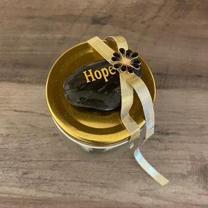 Könnte beinhalten: Ein Glasgefäß mit goldenem Deckel und einem schwarzen Stein, auf dem das Wort "Hope" eingraviert ist. Der Deckel ist mit einer schwarzen Blume und einem goldenen Band verziert.