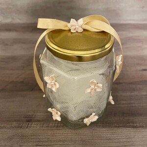 May include: A glass jar with a gold lid and a beige ribbon tied around the top. The jar is decorated with small, light pink fabric flowers and a white tulle fabric. The jar is filled with white glitter.