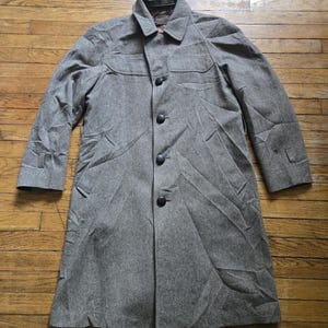 May include: A long, grey wool coat with a button-up front and a collared neckline. The coat has two front pockets and button details on the sleeves. The coat is laid flat on a wooden surface.