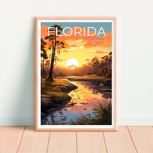 K&ouml;nnte beinhalten: Ein gerahmter Druck mit einer Sonnenuntergangsszene &uuml;ber einem Fluss in Florida. Der Druck zeigt einen leuchtend orangefarbenen und gelben Sonnenuntergang mit einer einzigen, gro&szlig;en, wei&szlig;en Sonne am Himmel. Der Fluss ist hellblau und auf beiden Seiten des Flusses befinden sich B&auml;ume. Der Text "FLORIDA" ist oben auf dem Druck in Wei&szlig; gedruckt.