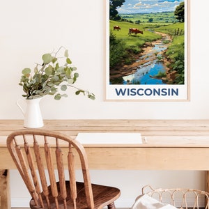 May include: A framed art print featuring a pastoral landscape with a stream, cows, and green fields under a blue sky. The word "WISCONSIN" is printed in navy blue at the bottom. The print is displayed on a wooden desk with a white vase and a wooden chair.