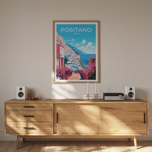 Może przedstawiać: Ramka z plakatem przedstawiającym kolorową ilustrację Positano we Włoszech. Plakat wisi na białej ścianie nad drewnianym regałem z dwoma głośnikami i systemem stereo.