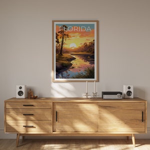 K&ouml;nnte beinhalten: Ein Holzschr&auml;nkchen mit drei Schubladen und einer Schiebet&uuml;r. Der Schrank steht in einem Raum mit einem gerahmten Poster, auf dem "FLORIDA" steht und ein Sonnenuntergang &uuml;ber einem Fluss abgebildet ist. Auf dem Schrank stehen zwei Lautsprecher und ein kleines silbernes Objekt in der Mitte.