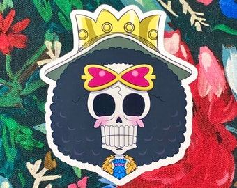 May include: A sticker featuring a cartoon skull with black curly hair, a yellow crown, and a pink bow tie. The skull has a wide grin and pink blush marks. The sticker is set against a floral background with red, pink, and green flowers.
