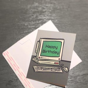May include: A birthday card featuring a retro computer design with a green screen displaying "Happy Birthday." The card is accompanied by a white envelope. The card's design includes a keyboard and mouse.