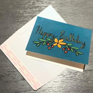 May include: A teal birthday card with the words "Happy Birthday" in gold script, adorned with a floral design. The card is accompanied by a white envelope, creating a thoughtful gift presentation.