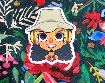 May include: A sticker featuring a cartoon character with tan skin, wearing a light green hat and headphones. The character has curly black hair and a small goatee. The background is a dark green fabric with a floral pattern in red, blue, and white.