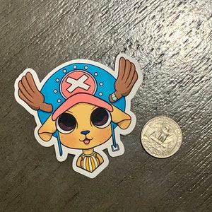 May include: A sticker featuring a cartoon character with a light yellow face, large black eyes, and a pink hat. The character has brown antlers and is set against a blue background with a white cross. A US quarter is next to the sticker.