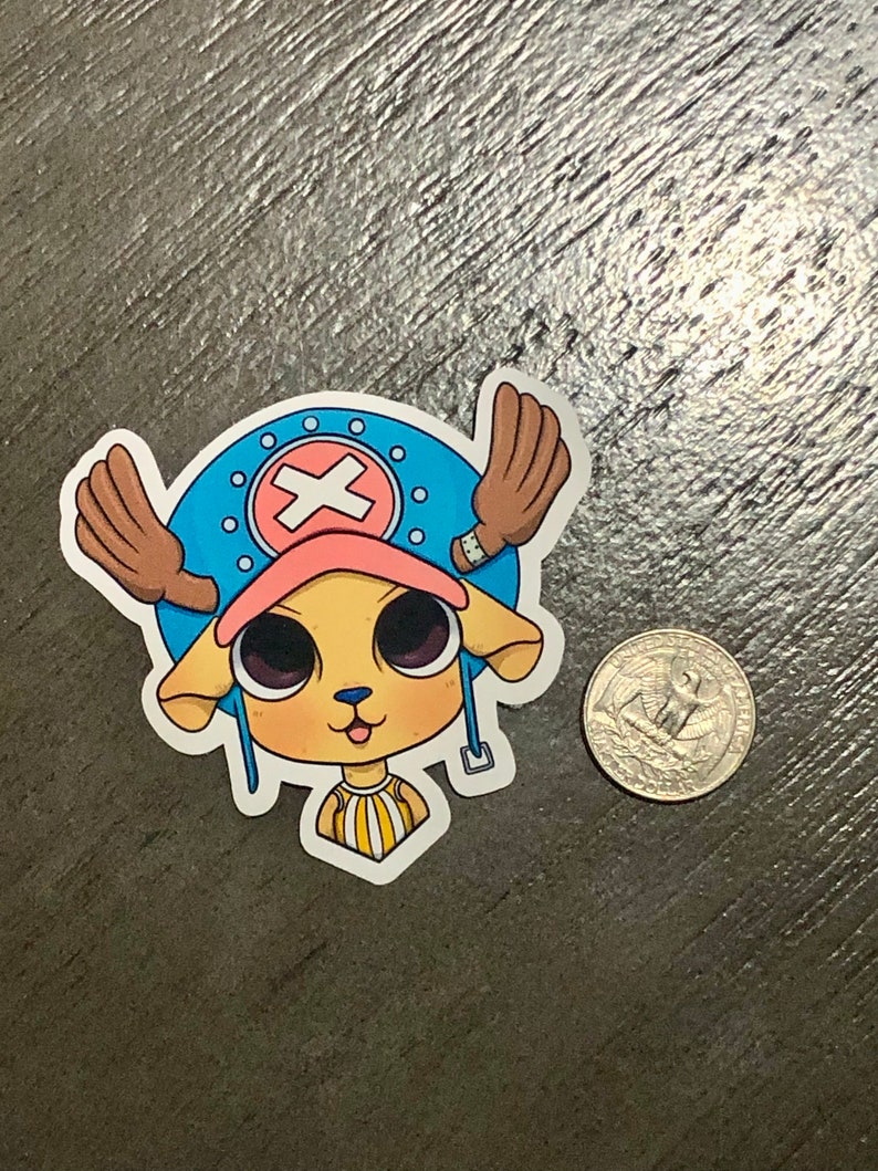 May include: A sticker featuring a cartoon character with a light yellow face, large black eyes, and a pink hat. The character has brown antlers and is set against a blue background with a white cross. A US quarter is next to the sticker.
