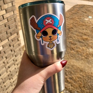 May include: A stainless steel tumbler with a sticker featuring a cartoon character with a blue hat, brown antlers, and a red cross symbol. The character has large eyes and a yellow and white striped collar.