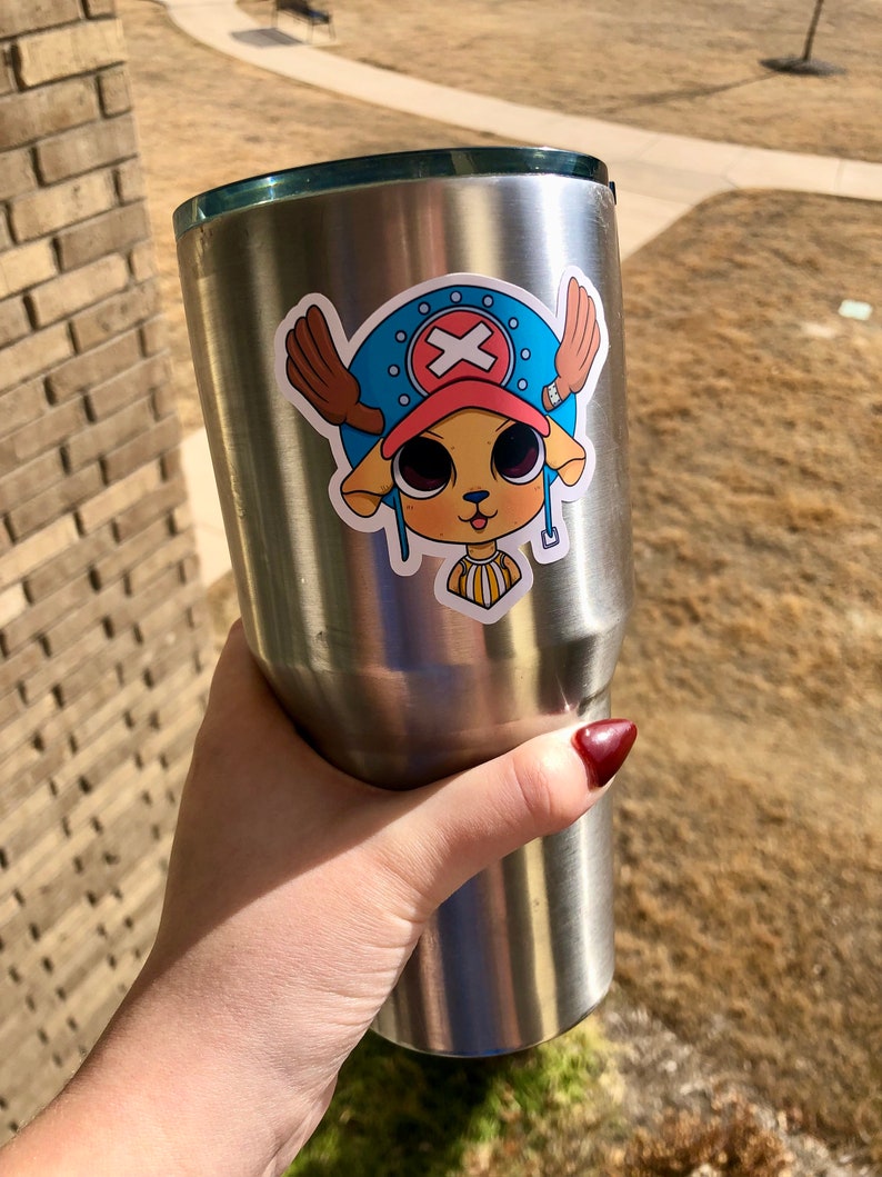 May include: A stainless steel tumbler with a sticker featuring a cartoon character with a blue hat, brown antlers, and a red cross symbol. The character has large eyes and a yellow and white striped collar.