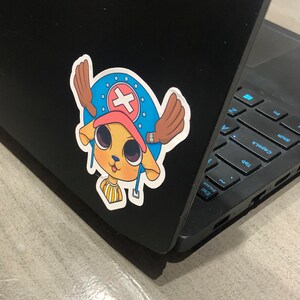 May include: A colorful sticker featuring a cartoon character with a blue hat, a red cross, and brown antlers. The sticker is affixed to a black laptop. The character has a yellow face and is smiling.