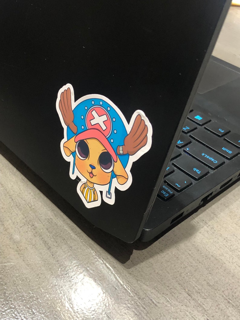 May include: A colorful sticker featuring a cartoon character with a blue hat, a red cross, and brown antlers. The sticker is affixed to a black laptop. The character has a yellow face and is smiling.