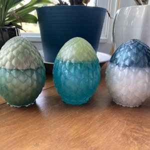 May include: Three decorative dragon eggs in shades of green, blue, and white. Each egg has a textured, scaled pattern and a two-tone color scheme. The eggs are displayed on a wooden surface.
