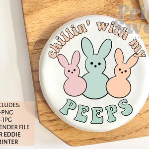 以下が含まれることがあります： 3匹のウサギのパステルグラフィックと「Chillin' with my Peeps」の文字が入った白い丸いクッキー。ウサギはピンク、ミントグリーン、ピーチ色です。クッキーは木製の表面にあります。画像にはファイル形式に関するテキストも含まれています。