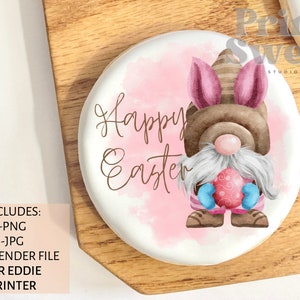 以下が含まれることがあります： ピンクの水彩画の背景と茶色の筆記体で「Happy Easter」と書かれた白い丸いクッキー。茶色の帽子、白いひげ、ピンクの耳をしたノームがピンクと白のイースターエッグを持っています。